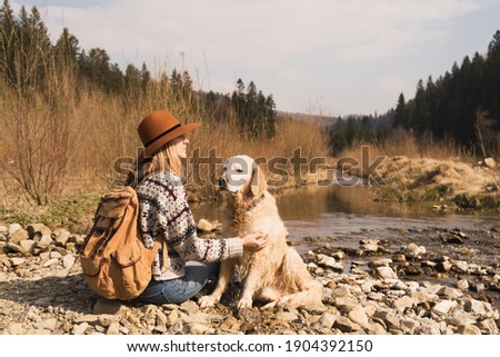 Traveling woman and golden retriever dog in nature. Stylish hipster in brown hat and wool sweater with vintage textile backpack near mountains river and forest.  Travel and wanderlust concept. 