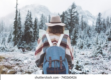Traveling woman standing in the wild with map in front of amazing view. Wearing hat, poncho and backpack. Winter is coming, first snowfall. Wanderlust and boho style