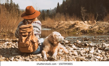 Traveling woman and wet golden retriever dog in nature. Stylish hipster in brown hat and wool sweater with vintage textile backpack near mountains river and forest.  Travel and wanderlust concept. 