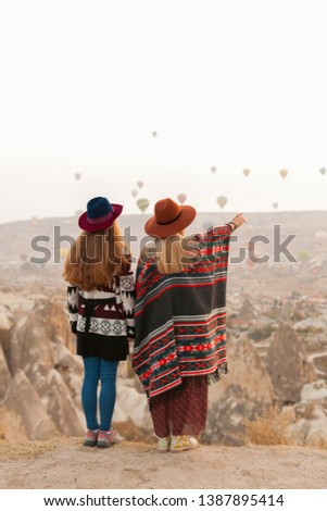 Traveling women wearing authentic boho chic style poncho, sweeter and hats looking on air ballons in sky in Cappadocia valley. Travel and wanderlust concept. Copy space background.