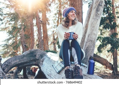 Traveling young woman with knitted hat drinking hot coffee from a mug and sitting on tree in wild forest at sunny day.  - Powered by Shutterstock