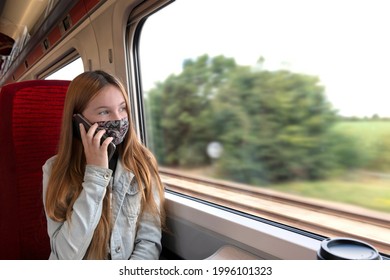 Travelling Female Teenager In Protective Mask Speaking On Mobile Phone While Sitting Near Window Inside Train During Summer Trip Through Countryside At Coronavirus Pandemic Perio