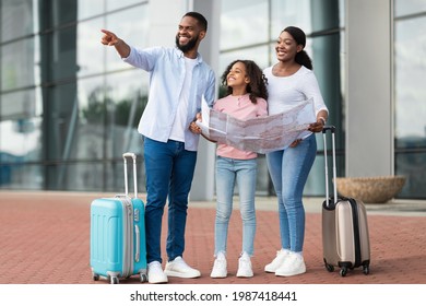 Travelling Together. Happy Young Black Family Standing Near Airport Building Holding City Map After Arrival To New Country, Cheerful People Planning Travel Route, Man Pointing Aside, Showing Way - Shutterstock ID 1987418441