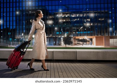 Travelling, tourism, business trip concept. Cheerful attractive mature lady wearing elegant outfit walking by airport at night, carrying her luggage, looking through window, copy space, full length - Powered by Shutterstock