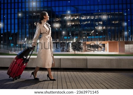 Travelling, tourism, business trip concept. Cheerful attractive mature lady wearing elegant outfit walking by airport at night, carrying her luggage, looking through window, copy space, full length