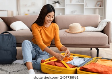 Travelling. tourism concept. Excited young asian woman sitting on floor in cozy living room, packing suitcase and backpack before journey at home, going vacation, copy space - Powered by Shutterstock