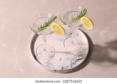 Tray with martini glasses of fresh cocktail, rosemary and lemon slices on beige marble table