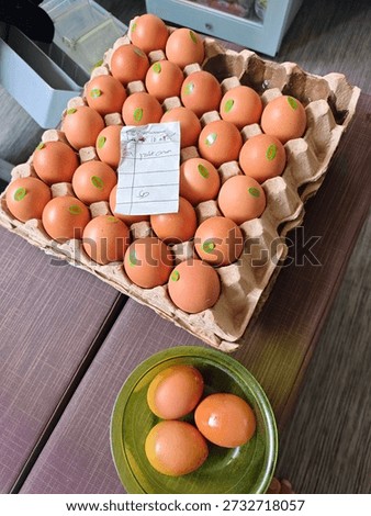 A tray of Omega-3 brown eggs, each marked with a small green sticker, sits neatly arranged in a cardboard carton, accompanied by a handwritten note reading “Yolk: Orange.” Below the tray, a green bowl holds four additional eggs, suggesting selection or inspection. Captured indoors in a modest kitchen or food storage space, with a refrigerator visible in the background, this image documents a quiet moment of food handling—possibly by a small-scale farmer, vendor, or home cook—who is taking pride in the quality and characteristics of their produce. The note emphasizes yolk color, hinting at the hens’ diet and the nutritional value of the eggs. The mood is intimate and observational, evoking care, authenticity, and a grassroots connection to food sourcing. It’s a visual narrative of trust, transparency, and the human touch behind everyday ingredients.