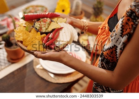 Tray with spicy chips, sauce, tacos and red hot chili peppers held by young Hispanic woman in national attire serving table for festive dinner