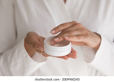 Treat yourself. Close up of millennial female wearing white bathrobe holding cosmetic cream jar. Young lady try moisturizing revitalizing creme lotion on finger before applying on hand face body skin