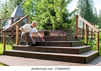 Treatment Of Wooden Boards With Protective Soil, Oil On Wood. A Man In A White T-shirt And Glasses 40-50 Years Old, Caucasian, Paints The Terrace. Painting A Wooden Terrace.