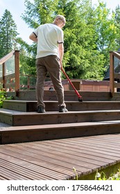 Treatment Of Wooden Boards With Protective Soil, Oil On Wood. A Man In A White T-shirt And Glasses 40-50 Years Old, Caucasian, Paints The Terrace. Painting A Wooden Terrace.