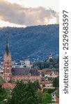 Tree branch framing distant view of majestic church Herz Jesu Kirche in Graz, Styria, Austria. Landmark against dramatic sky. Intricate architectural details, including pointed spire, ornate windows