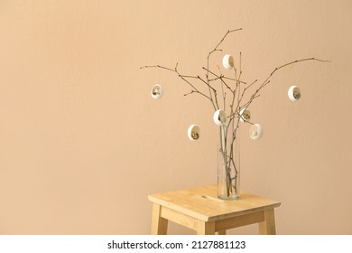 Tree branches decorated with cracked Easter eggs in vase on wooden stool near color wall