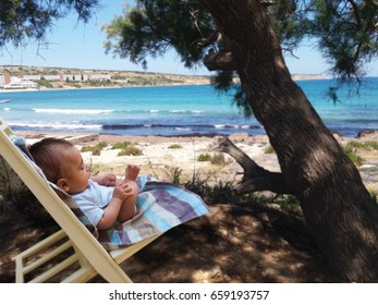 Tree Months Old Baby Boy Siting In Small Sun Bad Chair Relaxing In The Shadow Of The Tree On The Beach,Malta, Mellieha Bay