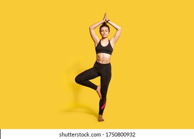 Tree pose. Athletic concentrated woman in tight sportswear practicing yoga, meditating with closed eyes, doing Vrksasana exercise on one leg, hands up. full length studio shot, sport workouts isolated