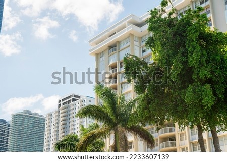 Trees at the front of condominium buildings in a row against the bright sky background in Miami, FL. Row of multi-storey residential buildings with balconies and modern architectural exterior.