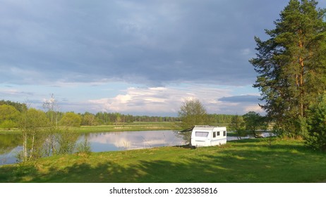 Trees grow on the grass-covered lawn on the river bank and there is a caravan trailer for convenience during fishing. There is a forest on the opposite bank. The river reflects the sky and clouds.