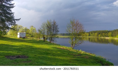 Trees grow on a lawn covered with grass on the river bank and there is a caravan trailer for comfort during fishing. There is a forest on the opposite bank. A thundercloud is coming.