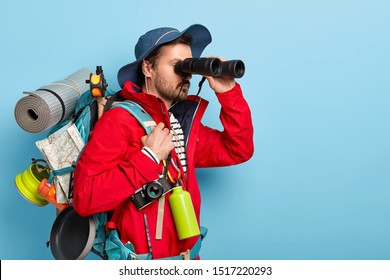 Trekking, camping, hiking concept. Photo of serious male tourist uses binoculars to observe surroundings, carries rucksack with rolled up rag, map and pan for cooking on bonfire, retro camera on neck