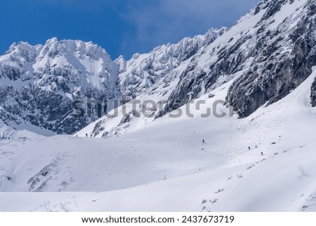Trekking and skituring in High Tatra Mountains in winter. Trail in snow with people and the giant rocky mountains in the bacground in sunny day with clear blue sky.