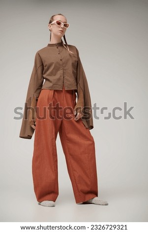 Trends. Haute couture collection. Beautiful fashion model girl posing in a designer brown shirt, knitted gloves and orange wide leg pants. Studio shot on a gray background. Stylish sunglasses. 商業照片 © 