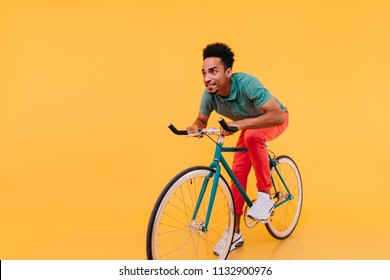 Trendy African Man In White Sneakers Posing Emotionally On Bike. Indoor Portrait Of Enthusiastic Sportive Guy Sitting On Bicycle.