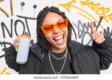 Trendy Afro American teenage girl smiles broadly makes yo gesture poses in urban place uses aerosol spray for drawing graffiti wears sunglasses and jacket belongs to hooligan gang. Youth subculture