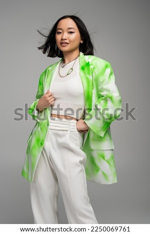 trendy asian woman in green and white blazer standing with hand on hip isolated on grey