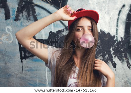 trendy beautiful long haired model posing on graffiti background. Blow bubblegum. red cap. grey t-shirt.