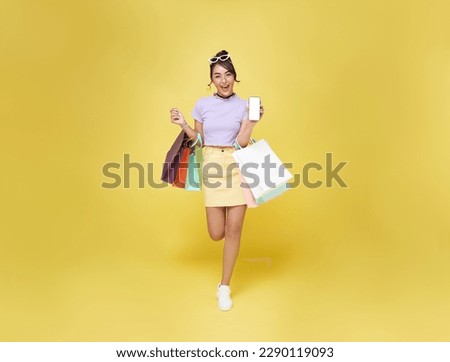 Trendy beautiful young Asian woman carrying shopping bags with showing mobile phone shopping online application isolated on yellow background.