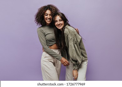 Trendy black girl in long sleeve top and wide modern pants smiling and hugging with dark haired lady in olive outfit on isolated backdrop.