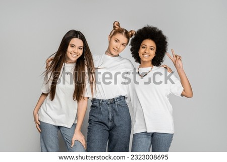 Trendy blonde teen girl in jeans and white t-shirt hugging multiethnic girlfriends and posing together isolated on grey, adolescence models and generation z concept, friendship and companionship