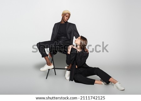 trendy blonde woman on chair and brunette woman on floor looking at each other on grey with shadow