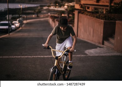 Trendy Boy Riding His Bicycle On The Warm Light Of A Summer Evening. Young Man Pedaling With BMX Alone On The Road Near The Sea. Teen That Fatigue Uphill On The Bike. Youth Recreation Positive Concept