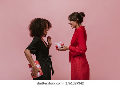 Trendy Brunette Lady With Silver Round Earrings In Red Bright Dress Giving Gift Her Curly Friend In Dark Clothes With Box.