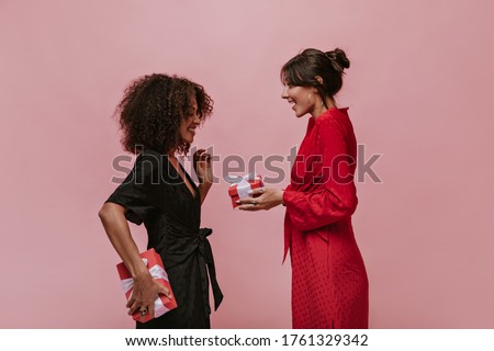 Trendy brunette lady with silver round earrings in red bright dress giving gift her curly friend in dark clothes with box.