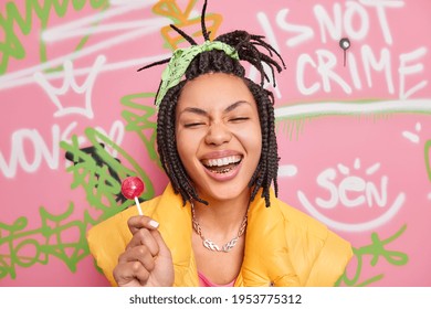 Trendy cheerful hipster girl smiles broadly holds lollipop has fun with teenagers of same age wears yellow vest poses against colorful graffiti wall. Gen z generation. Youth lifestyle concept - Powered by Shutterstock