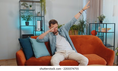 Trendy Cheerful Positive Adult Man Having Fun Dancing And Moving To Rhythm, Dabbing Raising Hands, Making Dub Dance Gesture. Young Caucasian Guy At Modern Home Apartment Living Room On Orange Couch