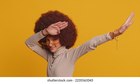 Trendy cheerful positive young school girl with afro hairstyle having fun dancing and moving to rhythm, dabbing raising hands, making dubdance gesture. Teenager female child kid on yellow background