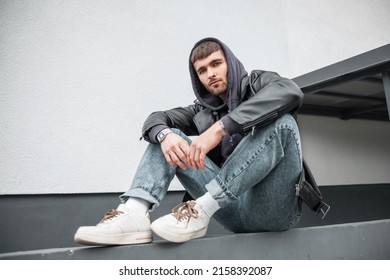 Trendy cool urban handsome man model hipster with leather jacket, hoodie, jeans and white sneakers sits on the street near a white wall - Shutterstock ID 2158392087