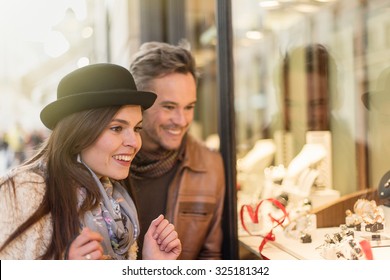 Trendy Couple Is Looking At Engagement Rings. A Grey Hair Man With Beard And A Woman With A Black Hat Are Standing In Front Of A Jewelry Shop. They Are Looking At The Shop Window.