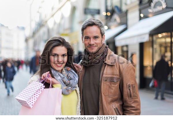 A trendy couple is standing arm in arm in the city\
center. They are in a cobbled car-free street. The woman is wearing\
a yellow shirt and pink shopping bags and the grey hair man has a\
leather coat
