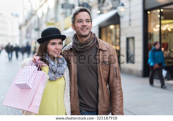 A trendy couple is walking arm in arm in the city\
center. They are in a cobbled car-free street. The woman is wearing\
a black hat and pink shopping bags and the grey hair man has a\
leather coat