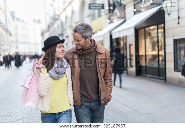 A trendy couple is walking arm in arm in the city
center. They are in a cobbled car-free street. The woman is wearing
a black hat and pink shopping bags and the grey hair man has a
leather coat