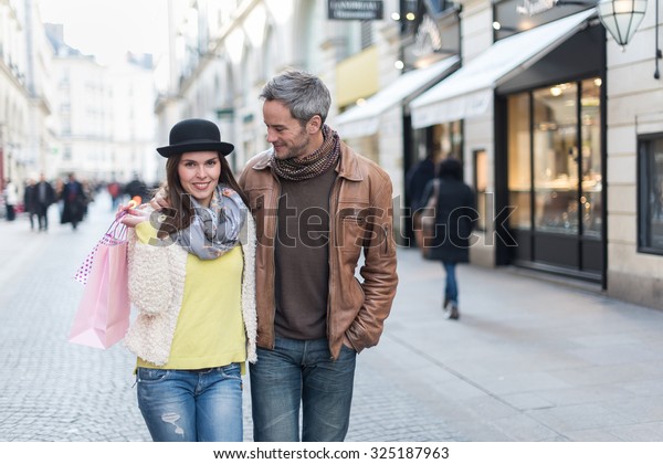 A trendy couple is walking arm in arm in the city\
center. They are in a cobbled car-free street. The woman is wearing\
a black hat and a pink shopping bag and the grey hair man has a\
leather coat