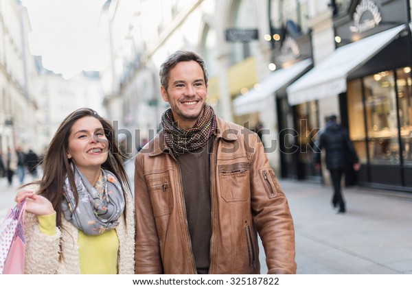 A trendy couple is walking in the city center. They\
are in a cobbled car-free street. The woman is wearing a yellow\
shirt and pink shopping bags and the grey hair man with beard has a\
leather coat