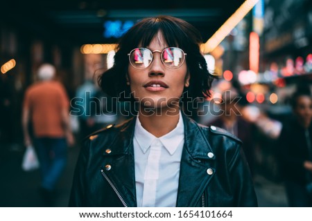 Trendy dressed tourist in stylish eyeglasses with neon reflection looking up during evening sightseeing around metropolitan downtown, fashionable woman in spectacles hanging out in night city