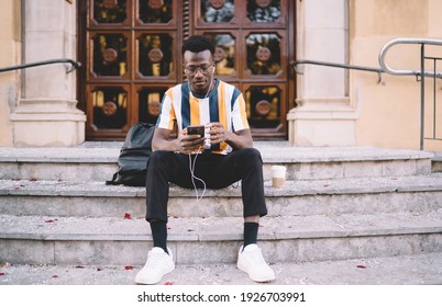 Trendy dressed travel blogger resting at street stairs using cellphone device for social networking, youthful hipster guy in spectacles and electronic headphones listening music and chatting