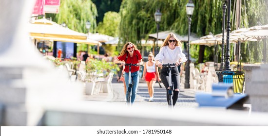 Trendy fashinable teenager girls riding public rental electric scooters in urban city environment. New eco-friendly modern public city transport in Ljubljana, Slovenia.